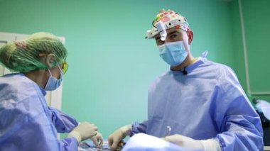 Farlı deneyimli bir erkek cerrah aletini değiştirip kameraya konuşuyor. Kendine güvenen doktor, kadın hemşirenin yardımıyla göz kapağı ameliyatı yapıyor..