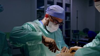 Gözlük takan erkek cerrah operasyon yerinde metal aletler kullanıyor. Doktora yardım etmek bir hastanın ayağını tutar..