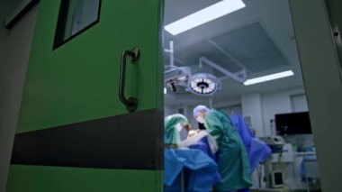 Modern bir ameliyathanenin kapısı kısmen açıldı. Ameliyathanede hastayı ameliyat eden iki doktor. Arkaplan bulanık. Düşük açı görünümü.