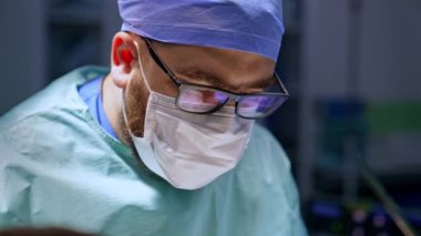 Gözlüklü, maskeli ve şapkalı beyaz erkek sakallı cerrah. Doktor ameliyata odaklanmış durumda. Kapat..