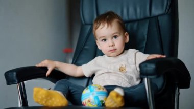 Güzel, beyaz bir bebek deri bir sandalyede topuyla oturuyor. Çocuk sakince oturur, sonra topu alır ve ayağa kalkar..