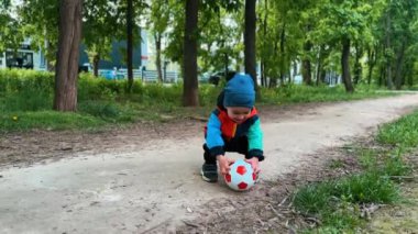 Şapkalı, pantolonlu ve ceketli sevimli çocuk topu yerden alıyor. Çocuk topu fırlatıyor yuvarlanışını izliyor ve neredeyse dengesini kaybediyordu..