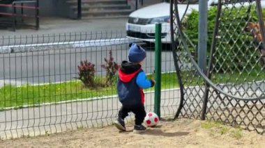 Kalın giysiler giyen çocuk dışarıda topla oynuyor. Çocuk yerden bir top alıyor ve onunla yürüyor..