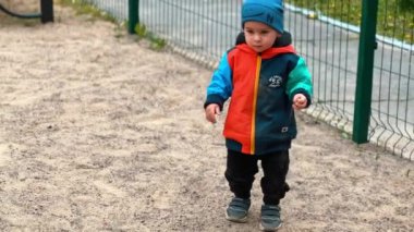 Beyaz çocuk oyun parkında kumla oynuyor. Çocuk avuç dolusu kumu alır ve hızla çitlere doğru fırlatır..
