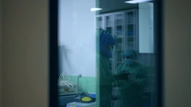 Ameliyata hazırlanan bir doktorun cam kapısındaki yansıma. Hemşire, cerrahın eldiven giyip bornozu bağlamasına yardım ediyor..