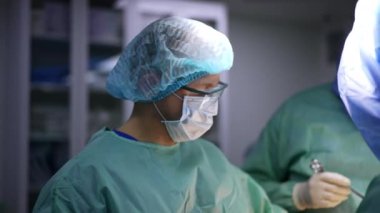 Gözlük, şapka ve maske takan erkek asistan cerrahi ekibinin yanında duruyor. Cerrahlara yardım eden doktor..