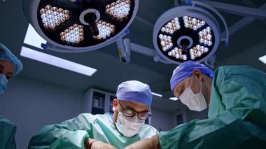 Ameliyat odasındaki yuvarlak lambaların altında iş birliği yapan cerrahlar. Hemşire doktorlara yardım ediyor. Düşük açı görünümü.