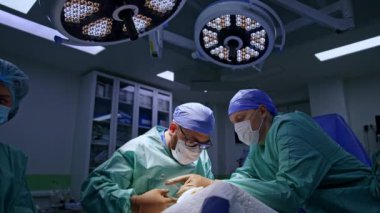 İki cerrah ameliyathanede yuvarlak lambaların altında çalışıyor. Hemşire enstrümanlarla masada duruyor..