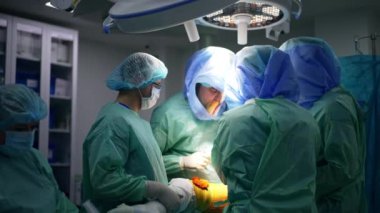Koruyucu giysi ve kask giyen profesyonel cerrahlar ameliyat masasının etrafında duruyorlar. Doktorlar bacağa artroskopi uygularlar..