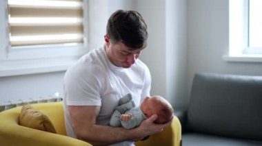 Beyaz tişörtlü beyaz bir adam elinde bebekle bir koltukta oturuyor. Yeni doğmuş baba, yeni doğmuş çocuğunu sakinleştiriyor..