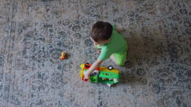 Bir yaşında bir çocuk odada halının üzerinde oturuyor. Siyah saçlı çocuk oyuncak trenle oynuyor. Üst görünüm.