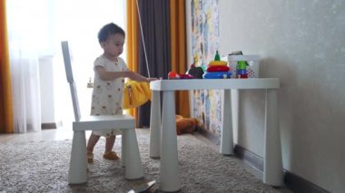 Küçük çocuk sandalyeleri masadan kaldırıyor. Bebek masadaki oyuncaklara dokunuyor ve sonra yere iniyor..