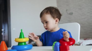 Mavi tişörtlü güzel erkek bebek masada oturuyor. Bir yaşındaki çocuk oyuncak piramidini monte ediyor..