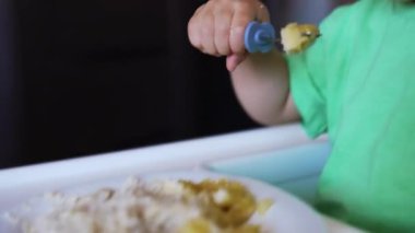 Yeşil tişört giyen tatlı erkek bebek çataldan yemek yiyor. Anne oğlunun yiyecek toplamasına yardım eder. Kapat..