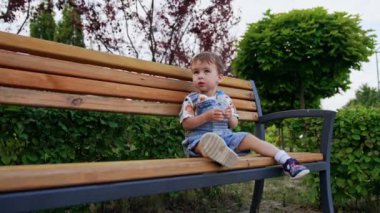 Yazın bankta oturan güzel beyaz bir bebek. Çocuk elinde bir şişe su tutuyor. Düşük açı görünümü.