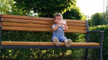 Parktaki bankta oturan sevimli erkek bebek. Çocuk şişeden su içiyor..