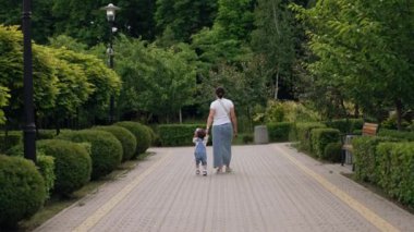 Koyu renk saçlı bir kadının bir bebekle el ele yürümesi. Anne ve oğlu yazın parkta yürüyorlar..
