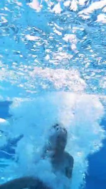 Çocuk açık mavi suya atlayıp bir sürü baloncuk yapıyor. Genç bir çocuk yüzme havuzuna atlıyor. Su altı kamerası. Dikey video