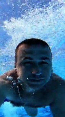 Yüzücü su altındaki kameraya bakarken ileri doğru yüzer. Yüzme havuzunda su altı kamerasıyla yüzen bir adam. Yaz tatili. Dikey video