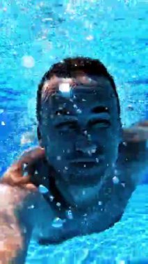 Adam yüzme havuzunda su altı kamerasıyla yüzüyor. Havuzun şeffaf su arka planı ve su altında yüzen ve kameraya bakan bir erkek. Dikey video