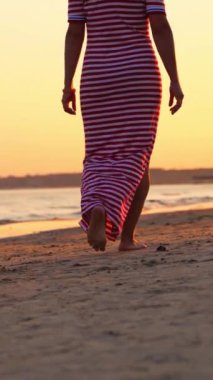 Gün batımında sahilde yürüyen bir kadının arka görüntüsü. Akşamları kumsalda çıplak ayakla yürüyen uzun kırmızı beyaz elbiseli genç bir kadın. Dikey video