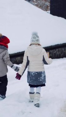 Çocuklar karlı evin yanında durur ve kışın konuşurlar. Dışarıdaki güzel beyaz karda yürüyen iki küçük çocuğun arka görüntüsü. Kalın elbiseli çocuklar Noel kartına bakarlar. Dikey video