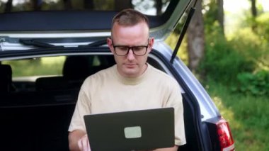 Orta yaşlı serbest yazar dizüstü bilgisayara odaklanmış. Adam ormanda bir arabanın bagajında çalışıyor..