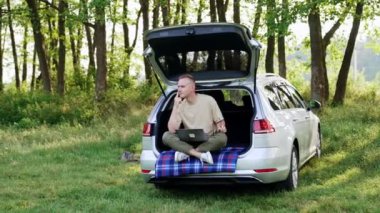 Düşünceli beyaz adam laptopuyla doğada çalışıyor. Mutlu adam arabanın bagajında oturur. Etrafına bakar, fikirleri düşünür..