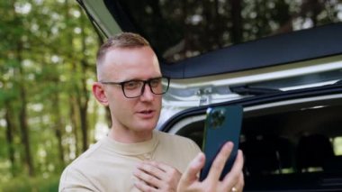 Gözlüklü beyaz erkek akıllı telefona bakıyor. Arabasının bagajında oturan adam telefonla konuşuyor ve saçlarını ekrandan kontrol ediyor..