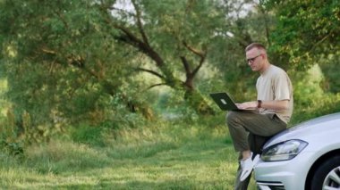 Beyaz bir erkek arabanın önünde dizüstü bilgisayar tutuyor. Bilgisayarda serbest çalışan adam tipleri ve gülümsemeleri..