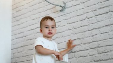 Beyaz duvarın yanında beyaz şirin beyaz bir çocuk duruyor. Bebek masadan bir şey almak için hızlı hareket ediyor..