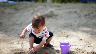 Hafif yaz kıyafetleri içindeki bebek eliyle biraz kum alıyor. Çocuk ayağa kalkar, kirli ellere bakar ve alkışlar..