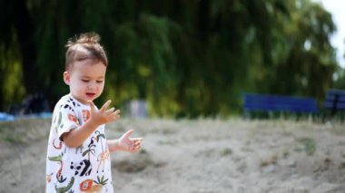 Tişörtlü güzel bir bebek güneşin altında dışarıda duruyor. Çocuk oyuncak küreği alır ve yere biraz kum toplar..