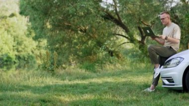 Gözlüklü orta yaşlı bir adam arabanın üstüne oturur. Serbest çalışan erkek, dizüstü bilgisayar ekranına bakıyor. Doğa zemin.