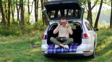 Arabasının bagajında oturan beyaz bir adam gözlük takıyor. Freelancer dizüstü bilgisayarı üzerinde çalışmaya hazırlanıyor. Güzel yaz doğası arka planda.