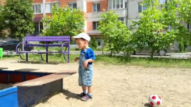 Şapkalı beyaz bir çocuk kum havuzuna doğru geliyor. Küçük çocuk kumla oynamak için içeri giriyor..