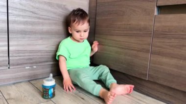 Güzel, beyaz bir bebek yerde rahatça oturuyor. Yeni yürümeye başlayan çocuk yere dokunur ve ayaklarını sallar. Çocuğun yanında emzik şişesi var..