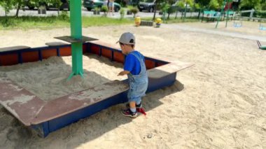 Kum havuzunun yanında kot pantolon ve kep giymiş güzel bir bebek oynuyor. Bebek daha fazla kum almak için dikkatlice içeri giriyor..