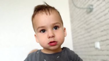 Gri tişört giyen iri gözlü, siyah saçlı, beyaz bir çocuk. Kameraya bakan sakin bir bebeğin yakın portresi. Beyaz arkaplan.