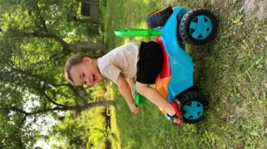 Güzel bir oyuncak arabanın üzerinde oturan gülen çocuk. Kafkasyalı çocuk yeşil çimlerin yanında tüm vücuduyla arabayı hareket ettiriyor. Dikey ekran.