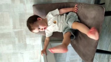 Beyaz erkek bebek yumuşak sandalyeye oturur ve küçük bacaklarını sallar. Anne küçük oğlunun ayaklarını gıdıklıyor. Dikey video.