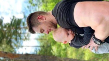 Sakallı beyaz adam ormanda kucağında küçük bir bebek tutuyor. Mutlu baba yeni doğmuş oğlunu öpüyor. Dikey ekran.