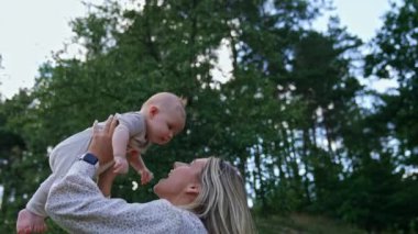 Sarışın, açık renk elbiseli bir kadın, küçük çocuğunu fırlatıyor. Mutlu kadın çocuğuyla doğada oynuyor..
