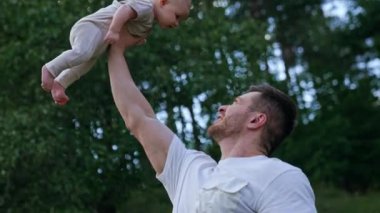 Kaslı beyaz bir adam tek eliyle yeni doğmuş bebeğini başının üstünde tutuyor. Gülümseyen mutlu baba oğlunu eğlendiriyor. Doğa zemin.