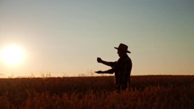Beline kadar çiftlikte duran bir adamın siluet profili. Şapkalı çiftçi ellerine tahıl döküyor. Arkaplanda gün batımı.