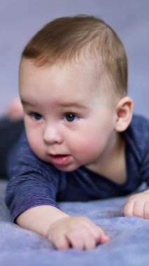 Güzel, tapılası erkek bebek yumuşak ekose üzerinde yatıyor. Küçük çocuk, ağzı açık ilgi çekici bir şeye bakıyor. Kapatın. Gri arka plan bulanık. Dikey video