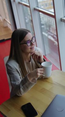 Masada çay içen uzun saçlı genç bir kadın. Yemek molasından hoşlanan ve pencereden dışarı bakan bir bayan. Yüksek açı. Dikey video