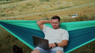 Neşeli beyaz adam hamakta dizlerinin üstünde dizüstü bilgisayarla oturuyor. Ofisten uzakta çalışan rahat bir adam..