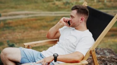 Sakin sakin adam beyaz tişört ve açık havada kot pantolon giyiyor. Adam telefonda konuşuyor rahat bir katlanır sandalyede oturuyor..