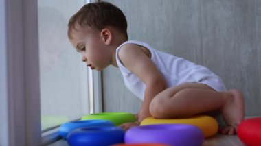 Sevimli beyaz çocuk panoramik pencerenin yanında yerde oturuyor. Çocuk önce aşağı bakar, sonra oyuncaklarına geri döner..